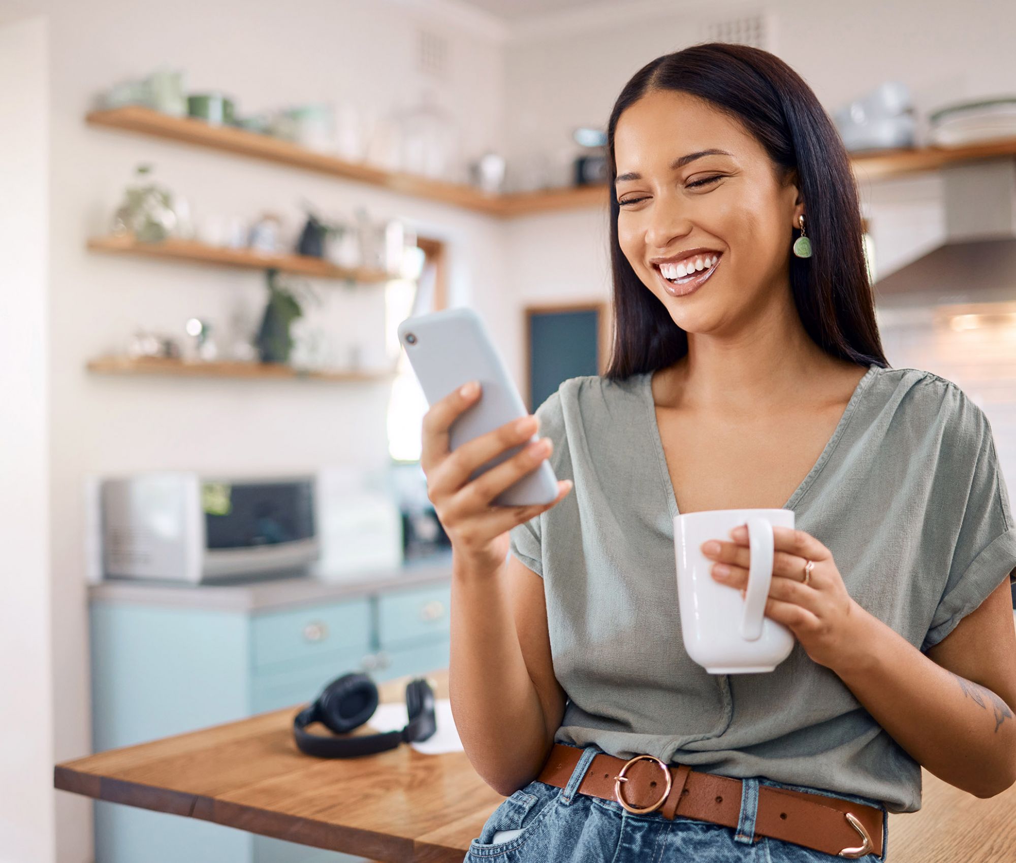 One happy young mixed race woman standing in her kitchen at home and using smartphone to browse the internet while drinking a cup of coffee. 