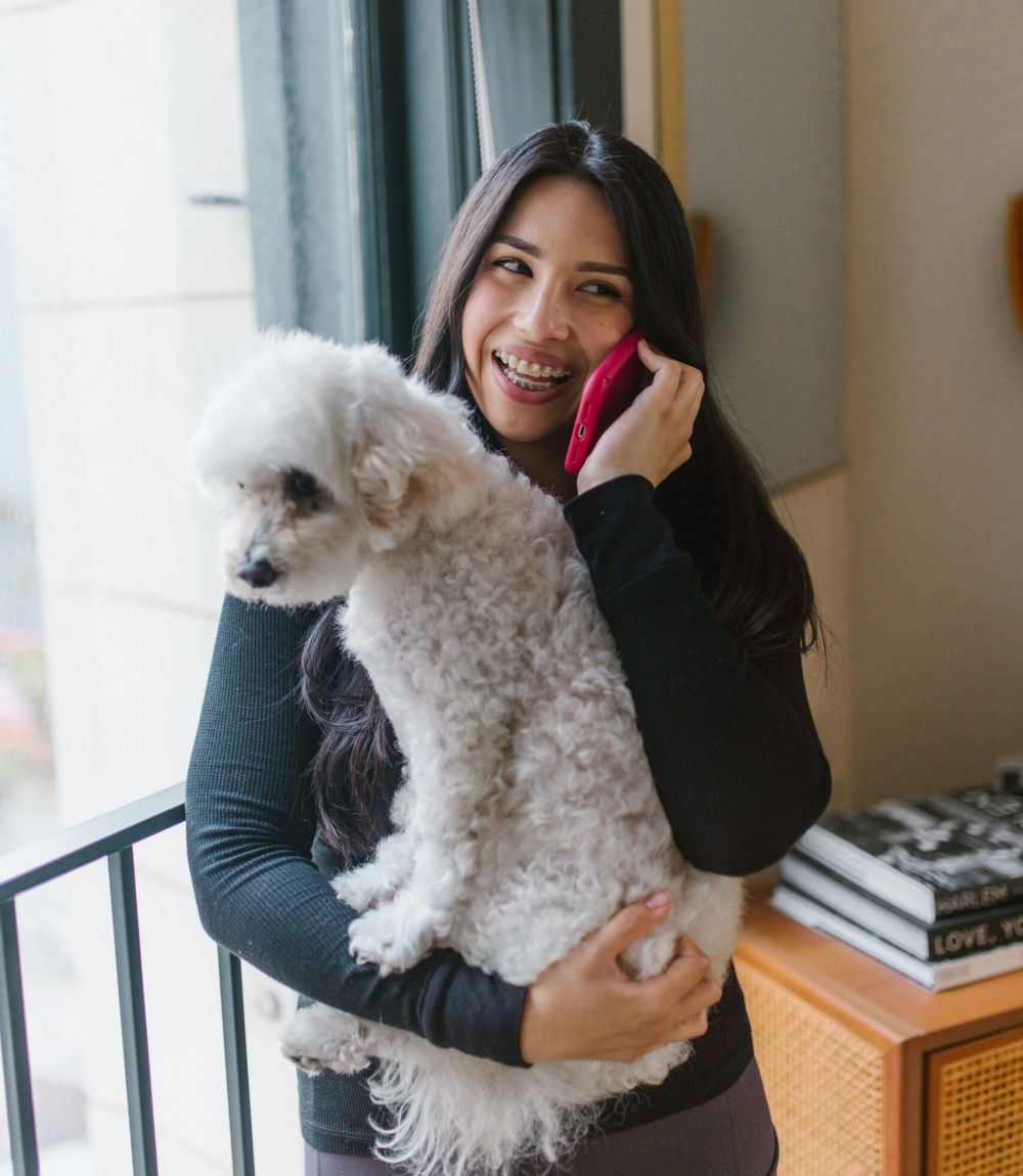 Smiling woman holding dog while talking on the phone in her apartment