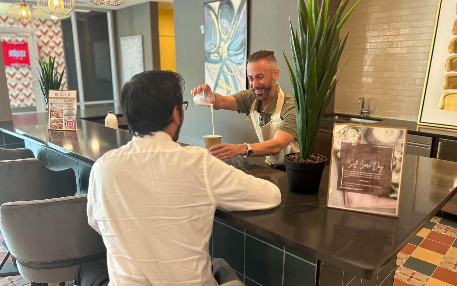 A woman and a man at a coffee counter, the woman is holding a coffee cup, both smiling. Art and decor in the background.